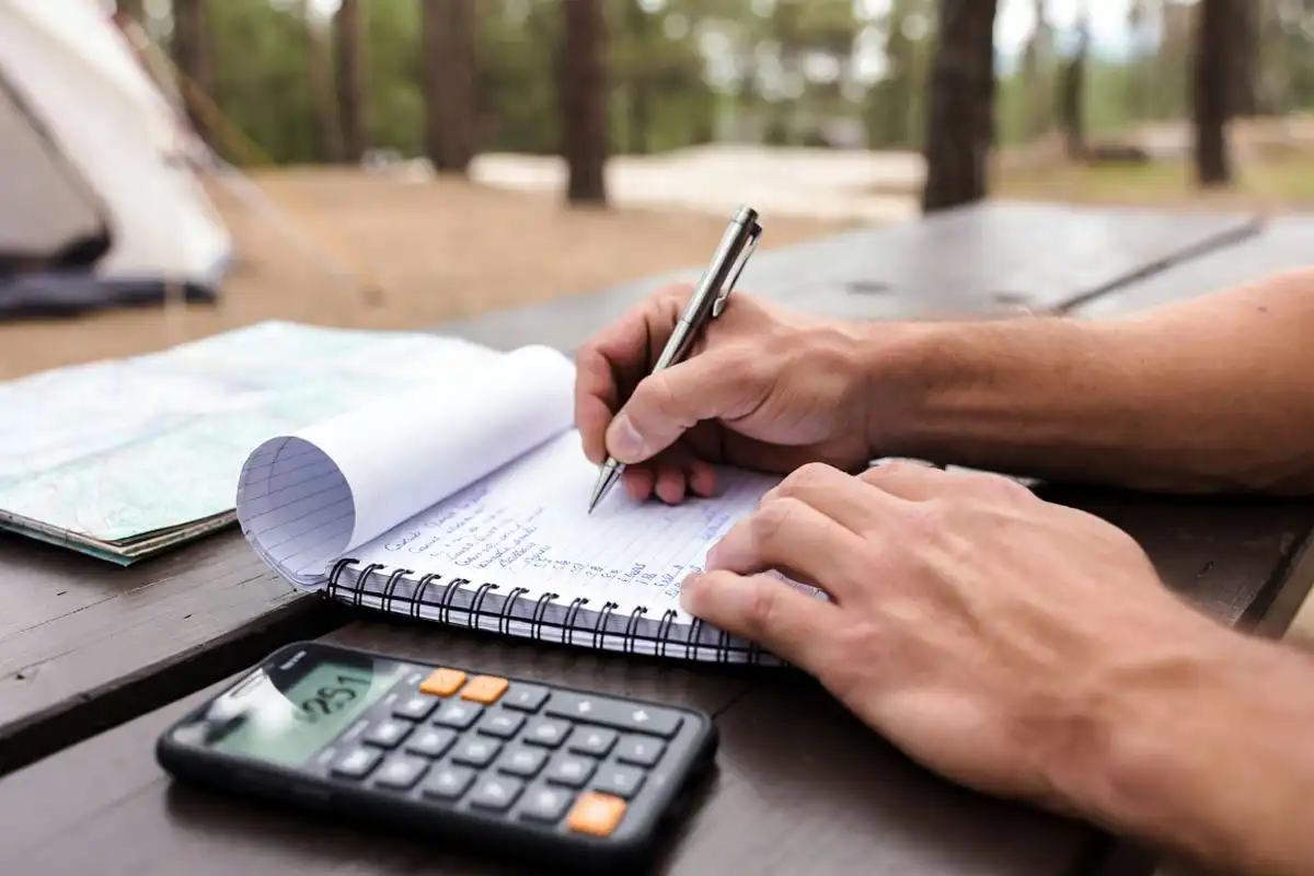 man writing camping cost breakdown on notepad at picnic table with map beside him