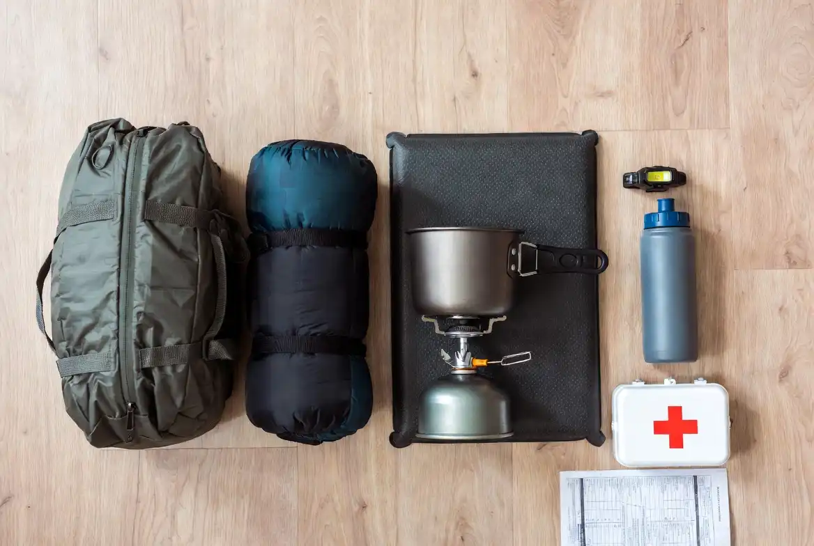 flat lay of camping essentials including tent, sleeping bag, headlamp, and camp stove