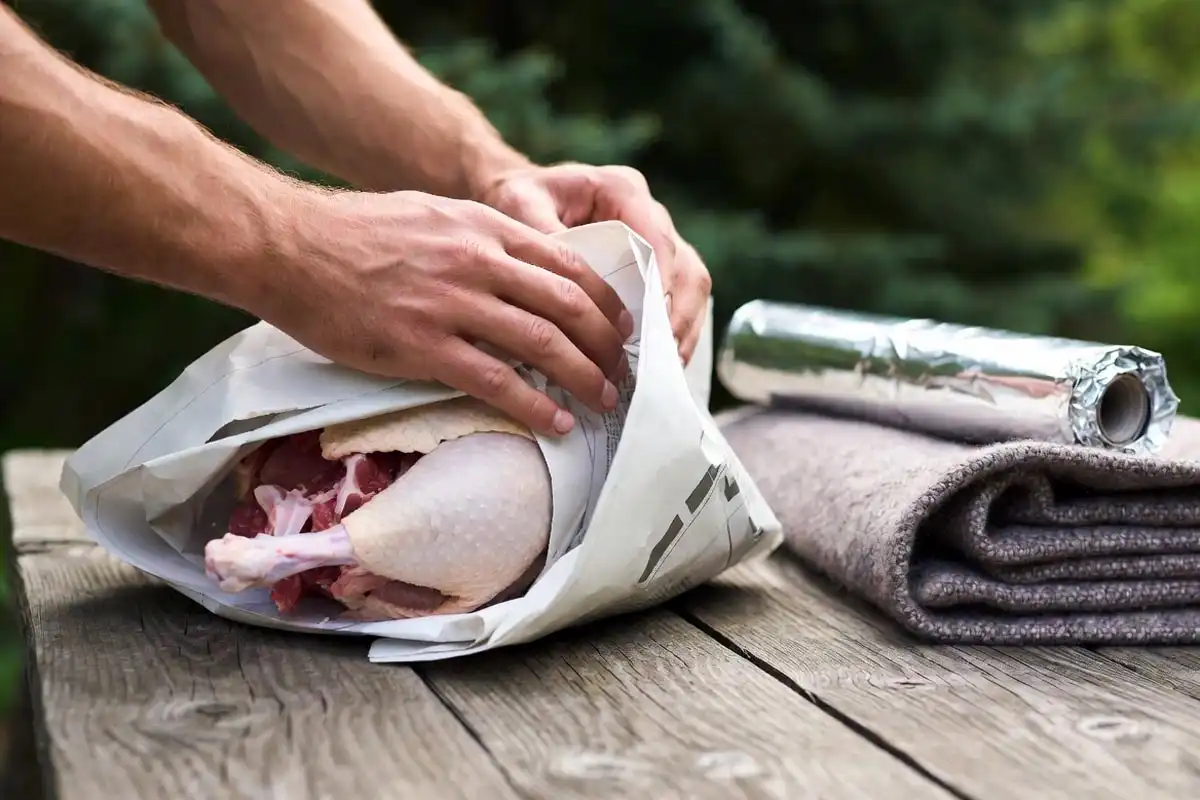 Camper wrapping food items in thick newspaper layers on a wooden camp table