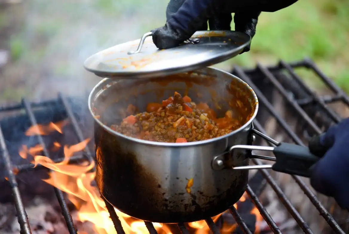 camper lifting lid of simmering camp pot with vegetable stew over outdoor stove