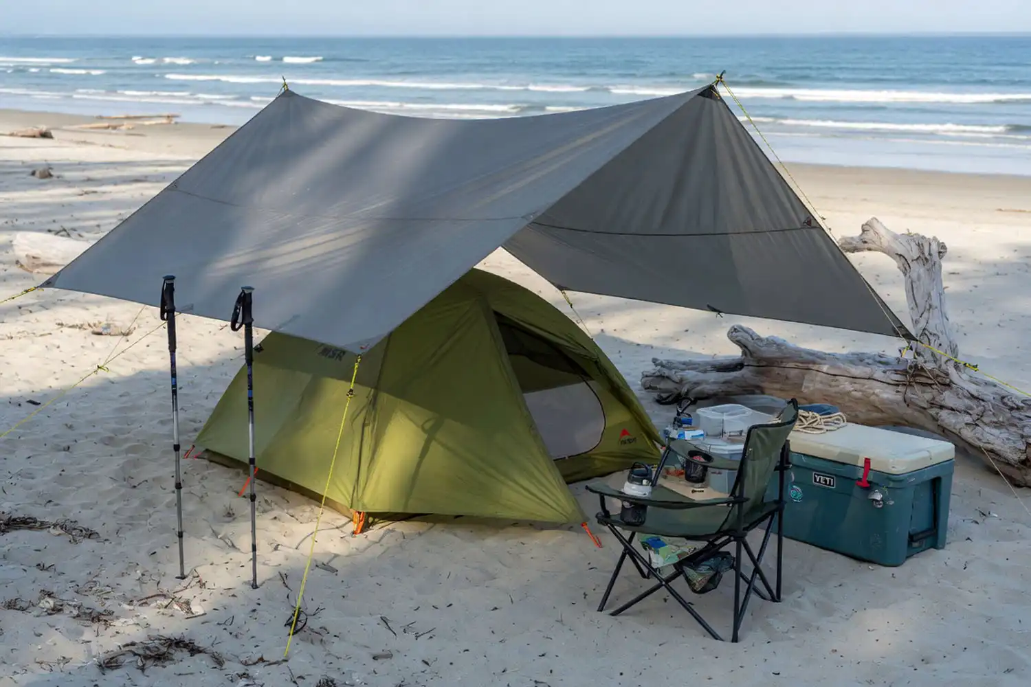 Beach campsite with a tarp angled for shade over a tent next to a cooler and camp chair on a sunny coastal afternoon