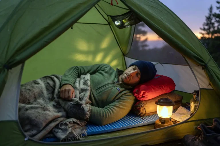 Camper resting on a foam pad under a wool blanket inside a small tent at dusk without a sleeping bag