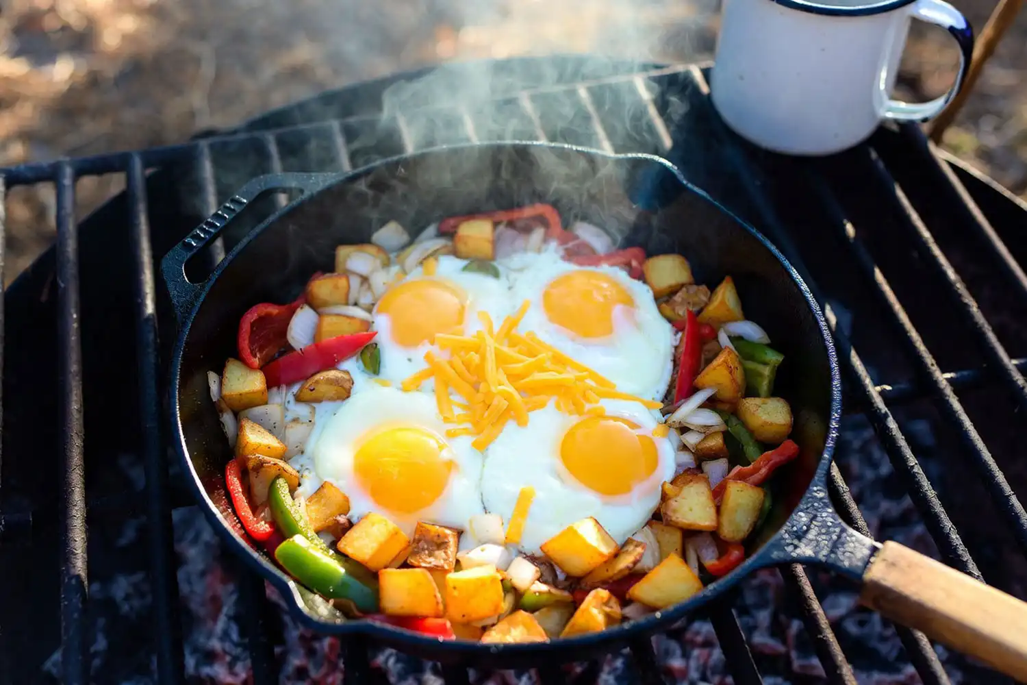 Cast iron skillet of potato hash with red and green bell peppers onions and four sunny side up eggs cooking on a campfire grate in the morning