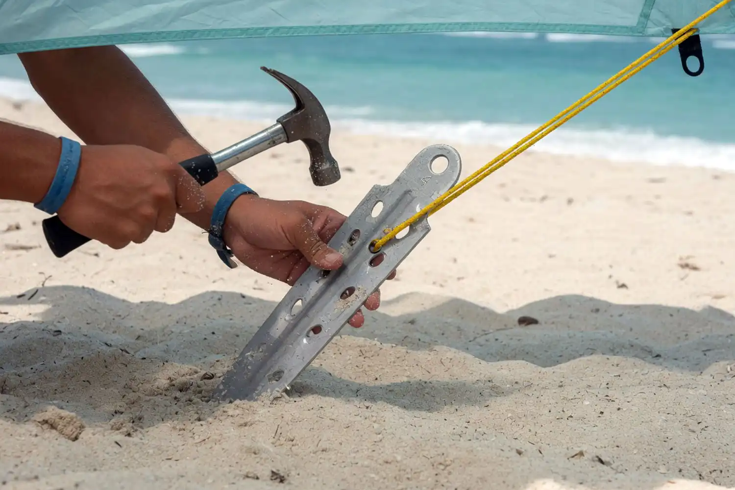 Camper using a wide aluminum sand stake to anchor a tent guy line on a sandy beach with the ocean in the background