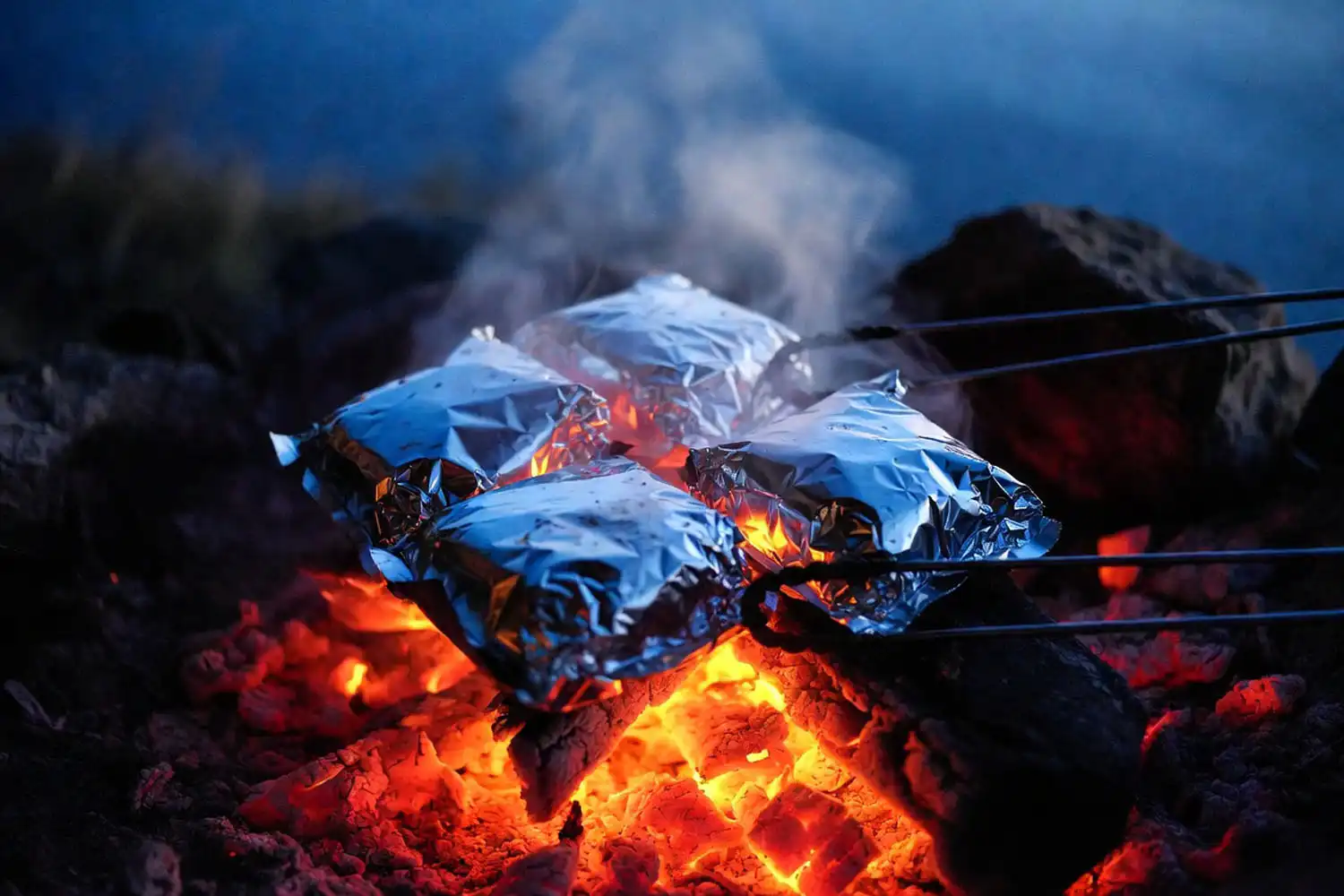 Four sealed aluminum foil packet dinners cooking on glowing orange campfire coals with tongs resting beside them