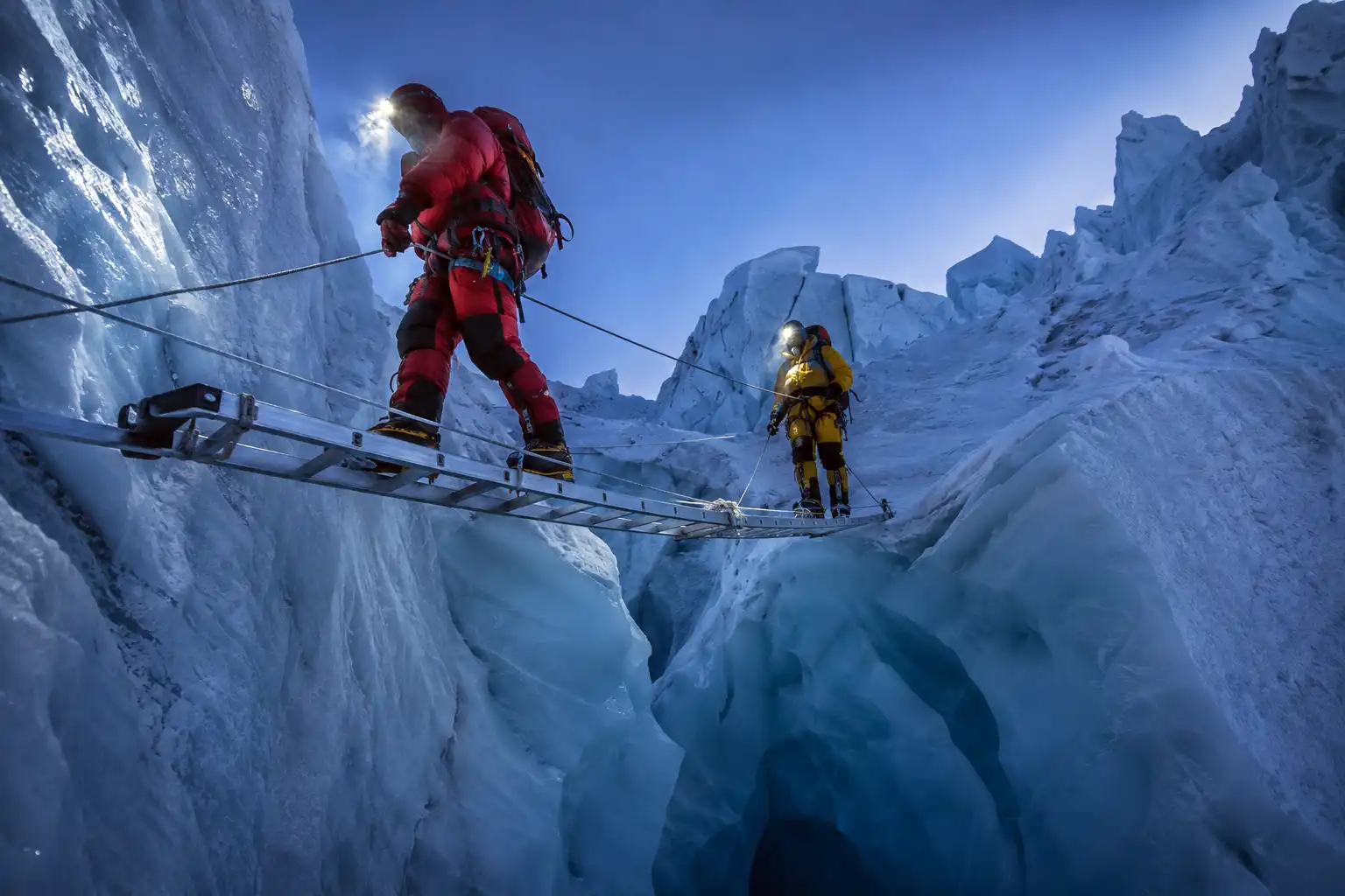 Climbers crossing an aluminum ladder over a deep blue crevasse in the Khumbu Icefall below Camp 1 on Mount Everest