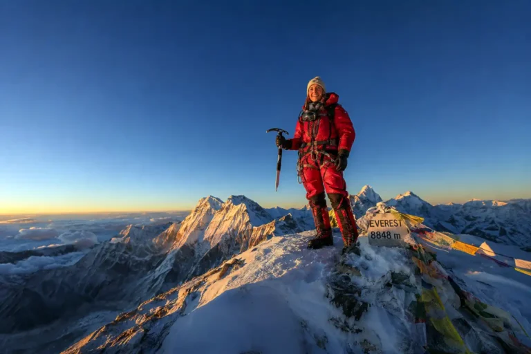 Female mountaineer reaching the summit ridge of Mount Everest at sunrise with prayer flags visible