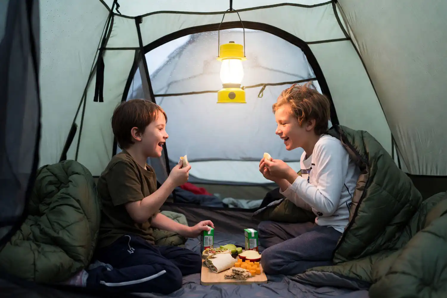 Kids eating no-cook camping meals of wraps, fruit, cheese, and granola bars inside a tent while rain falls outside