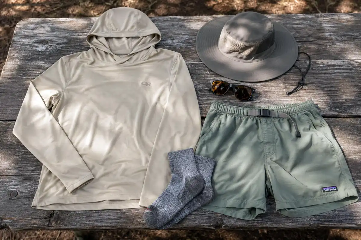 Lightweight summer camping clothes including a sun hoody, hiking shorts, and merino socks laid out on a wooden picnic table at a forest campsite