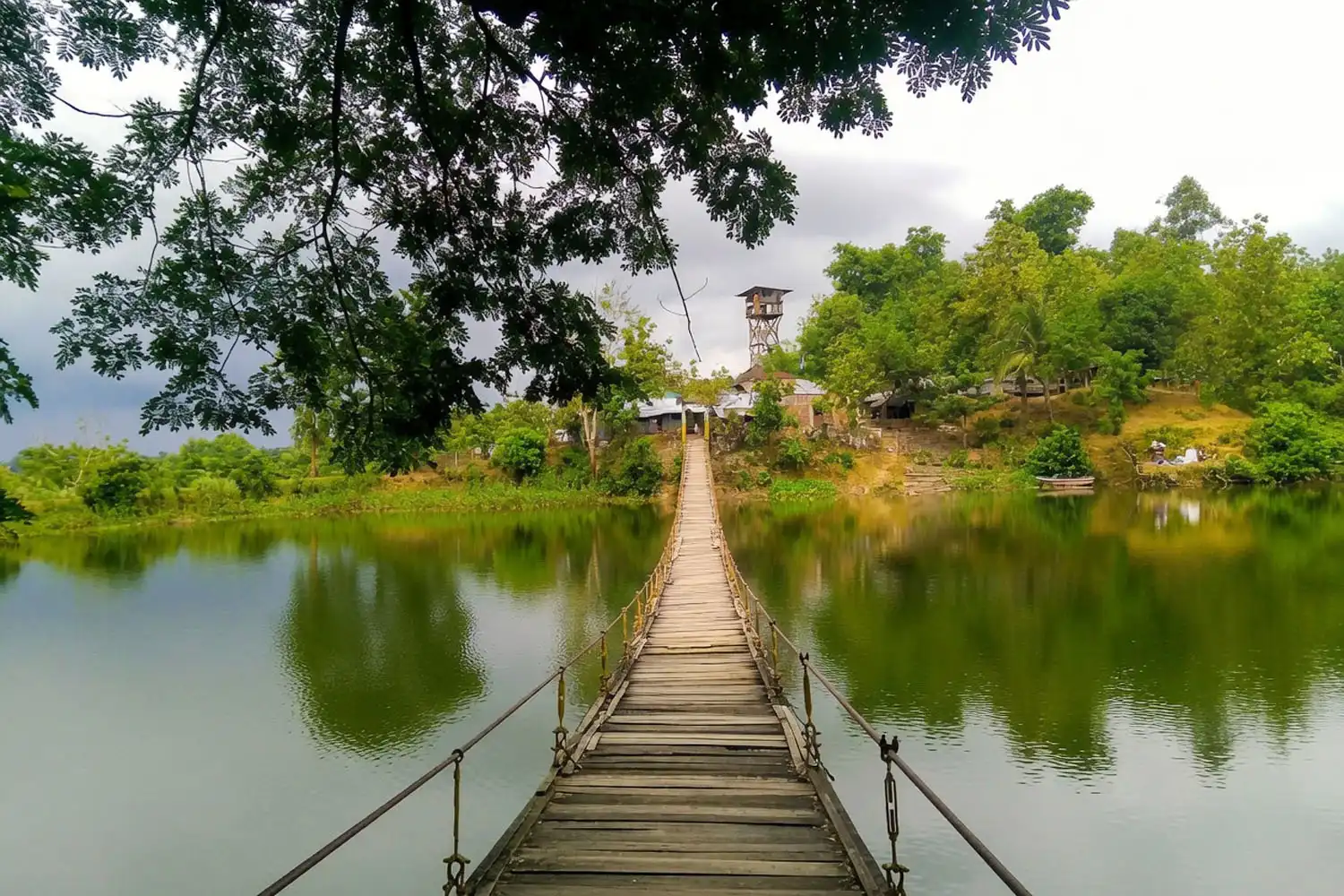 Hanging bridge crossing the green water of Naikhongchhari Lake with a watch tower and forested hills behind it