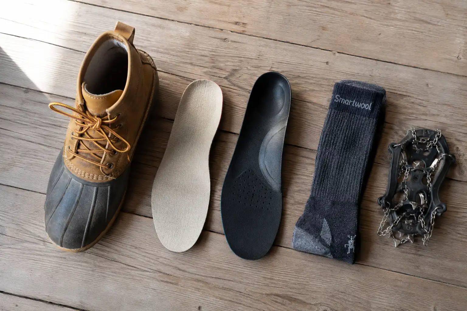 Flat lay of a duck boot beside a firm aftermarket insole with a deep heel cup, a folded merino wool sock and a pair of microspikes