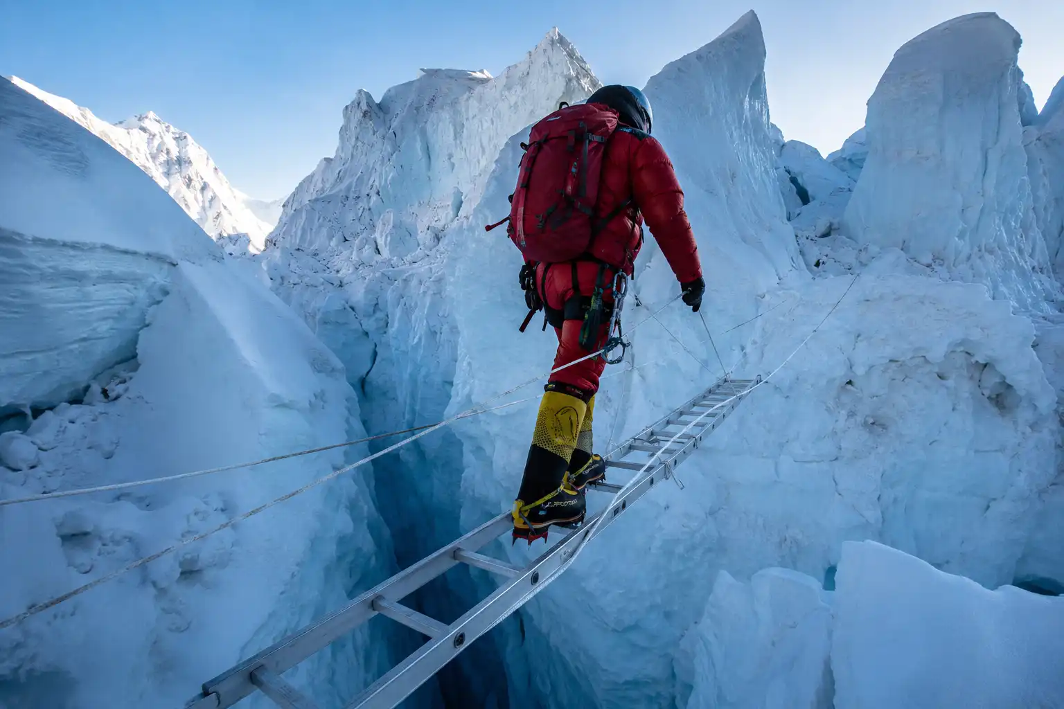 Roped climber in a red down suit crossing an aluminum ladder over a deep crevasse in the Khumbu Icefall on Mount Everest