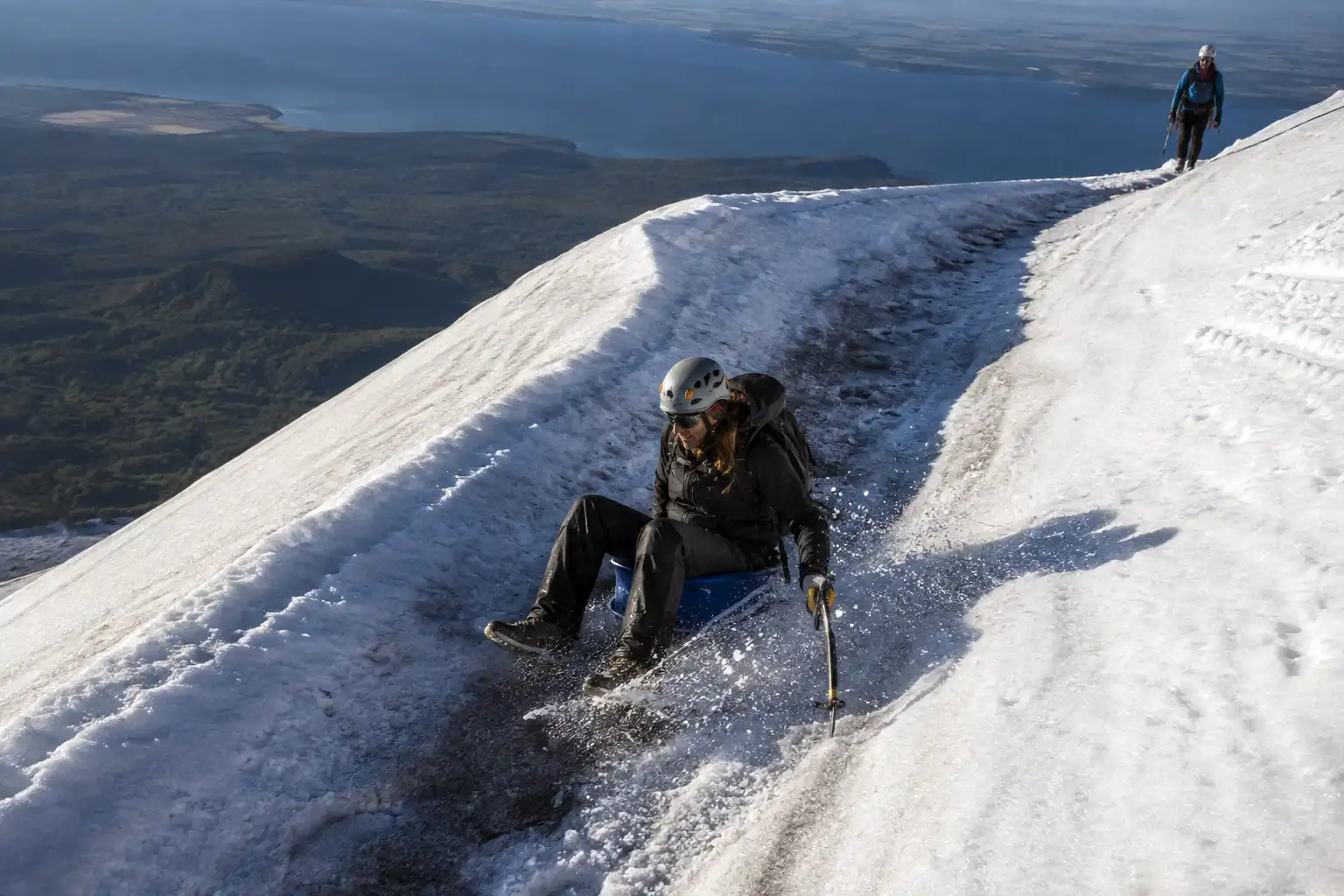 Climber in waterproof over-trousers sliding down a groove in the snow on a plastic sled while descending Villarrica volcano