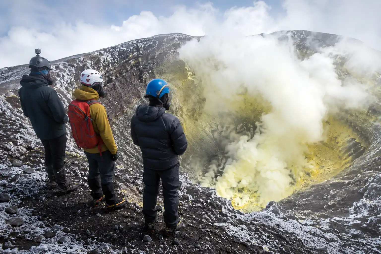 Climbers in helmets and gas masks standing at a safe distance from the steaming open crater at the summit of Villarrica volcano