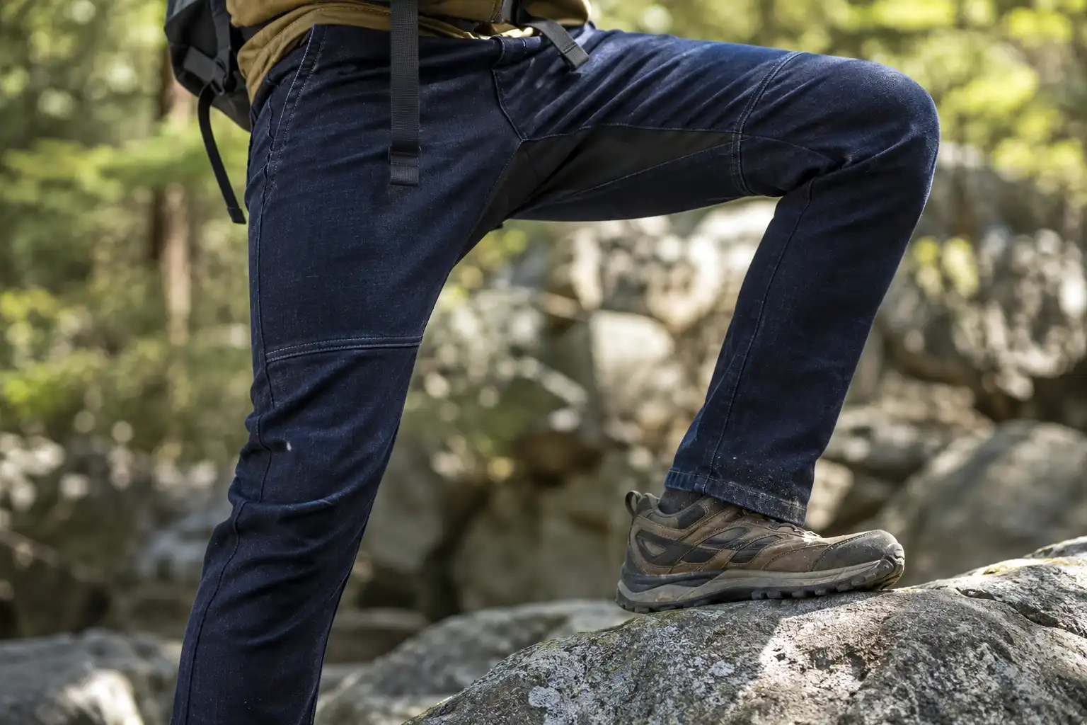 Close up of a hiker's leg in stretch performance denim bending sharply at the knee on a rock step, showing the fabric flexing without pulling tight