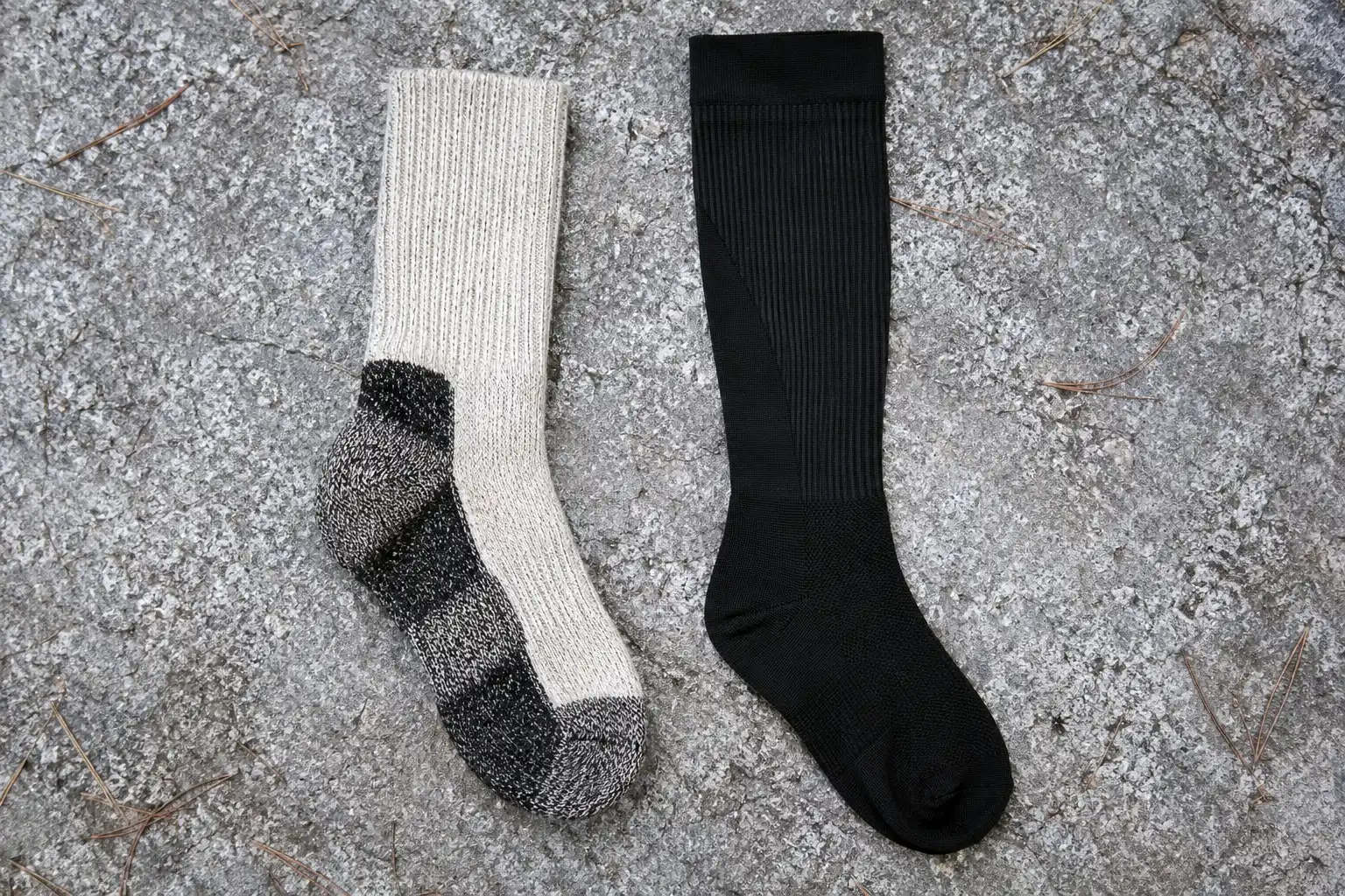 Thick cushioned merino hiking sock laid beside a thin knee-high compression sock on a flat rock to compare thickness and length