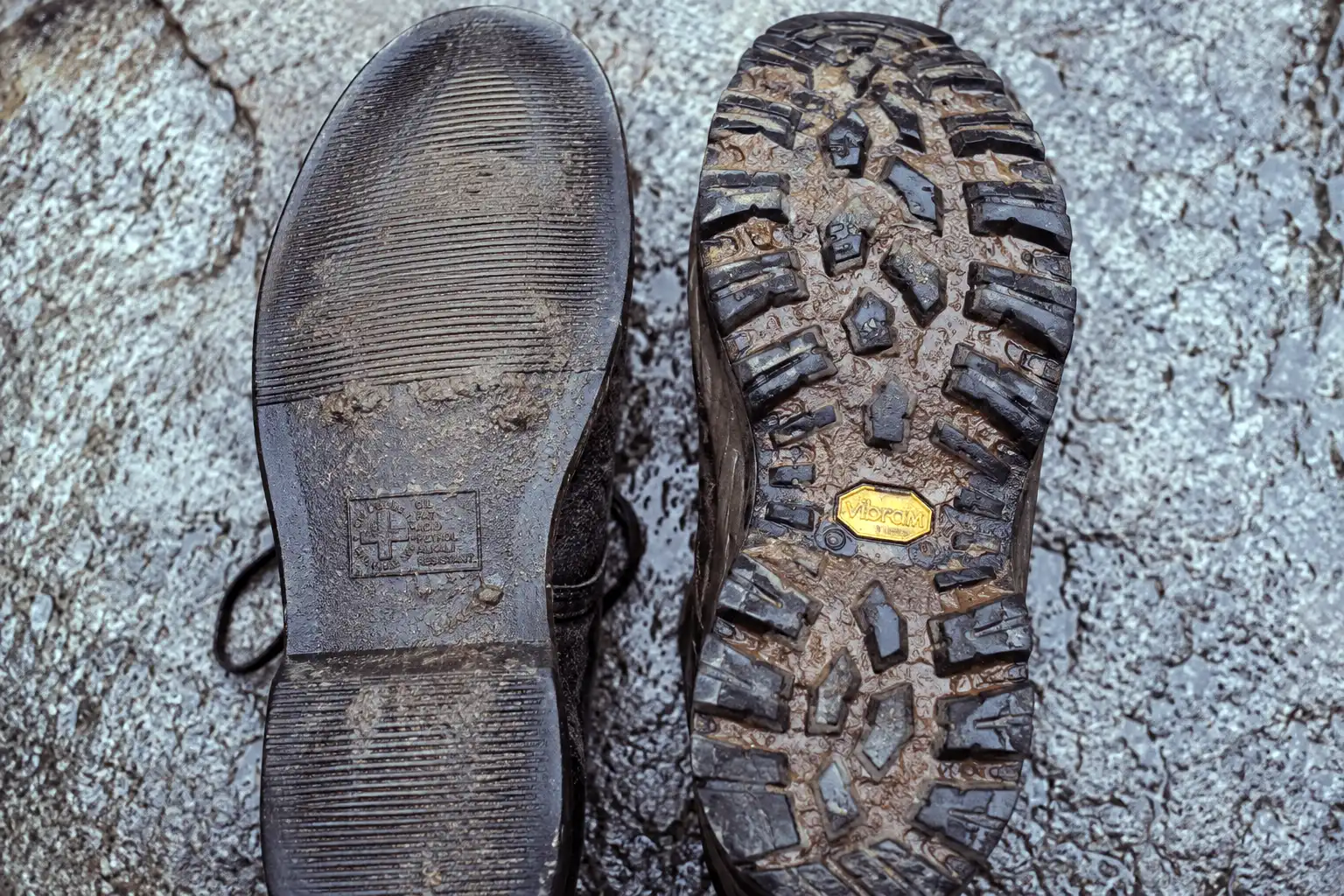 Close-up of a flat shallow-grooved Doc Martens sole next to a deep-lugged hiking boot outsole caked with mud on a wet stone surface