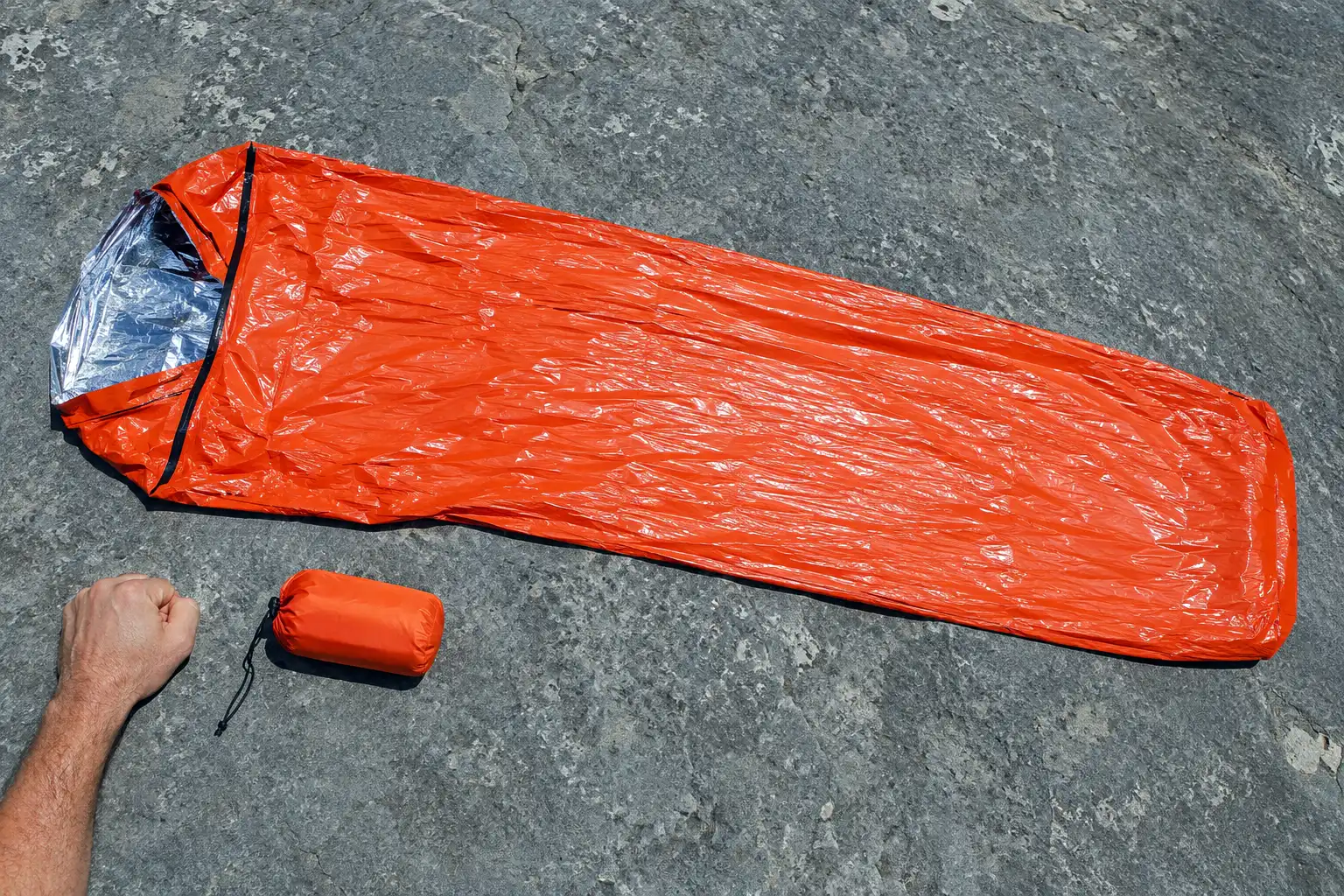 Bright orange single use emergency bivy sack unrolled on rock next to the fist sized stuff sack it packs into, showing how little space it takes