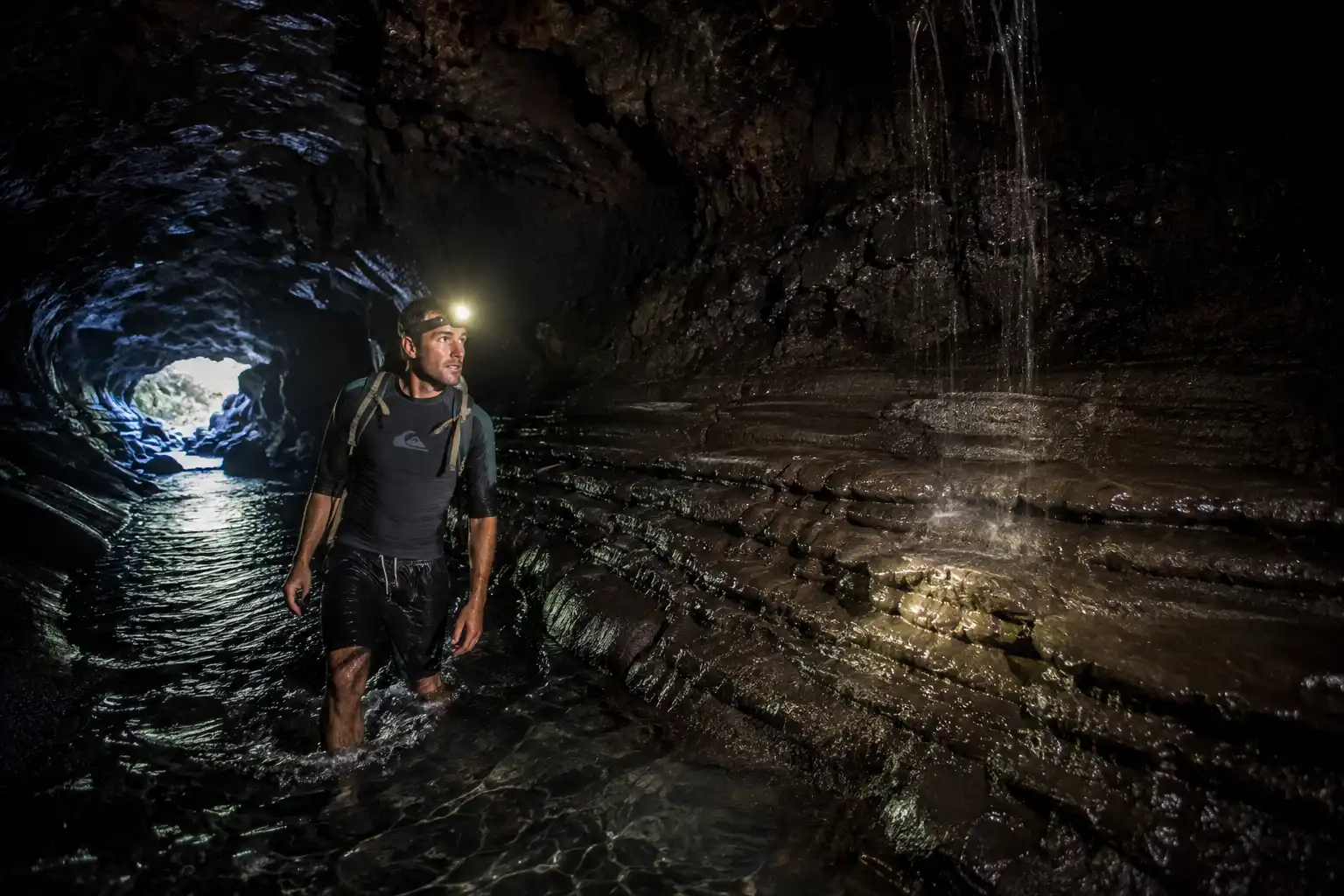 Hiker wading through knee deep water in a dark lava tunnel lit only by a headlamp