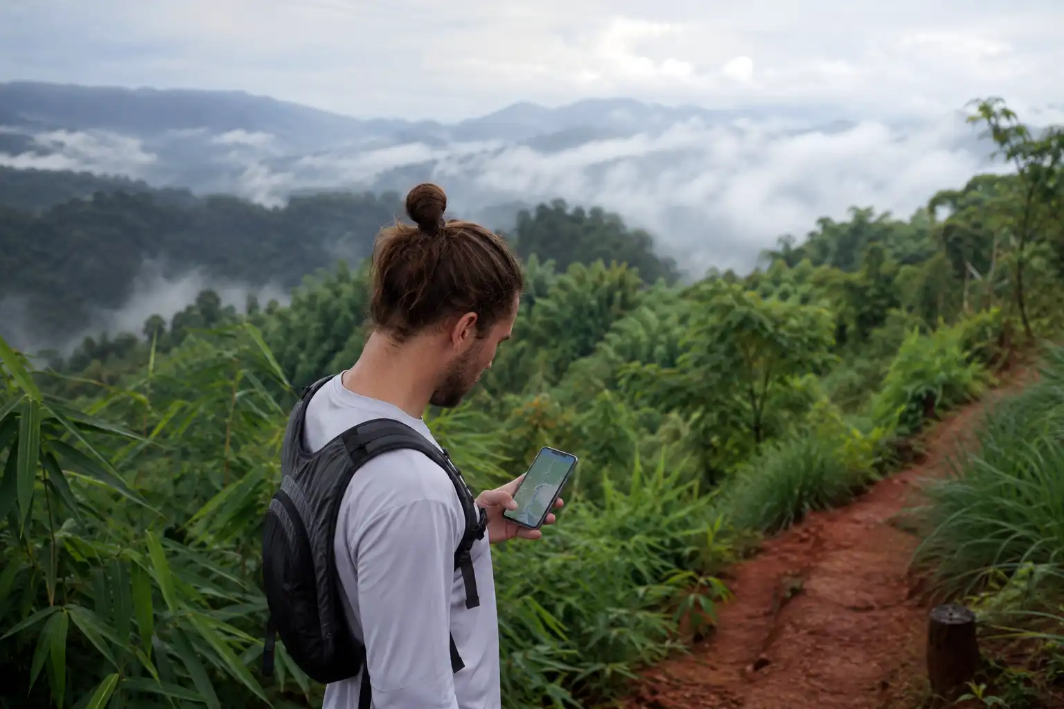Hiker pausing on a green hill path in the Chittagong Hill Tracts to check route notes on a phone before timing a run