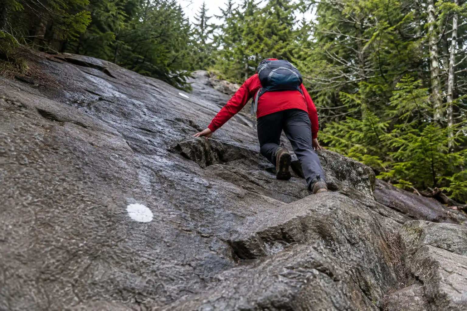 Hiker using hands to climb a steep section of bare granite slab on the White Dot Trail below the summit of Mount Monadnock