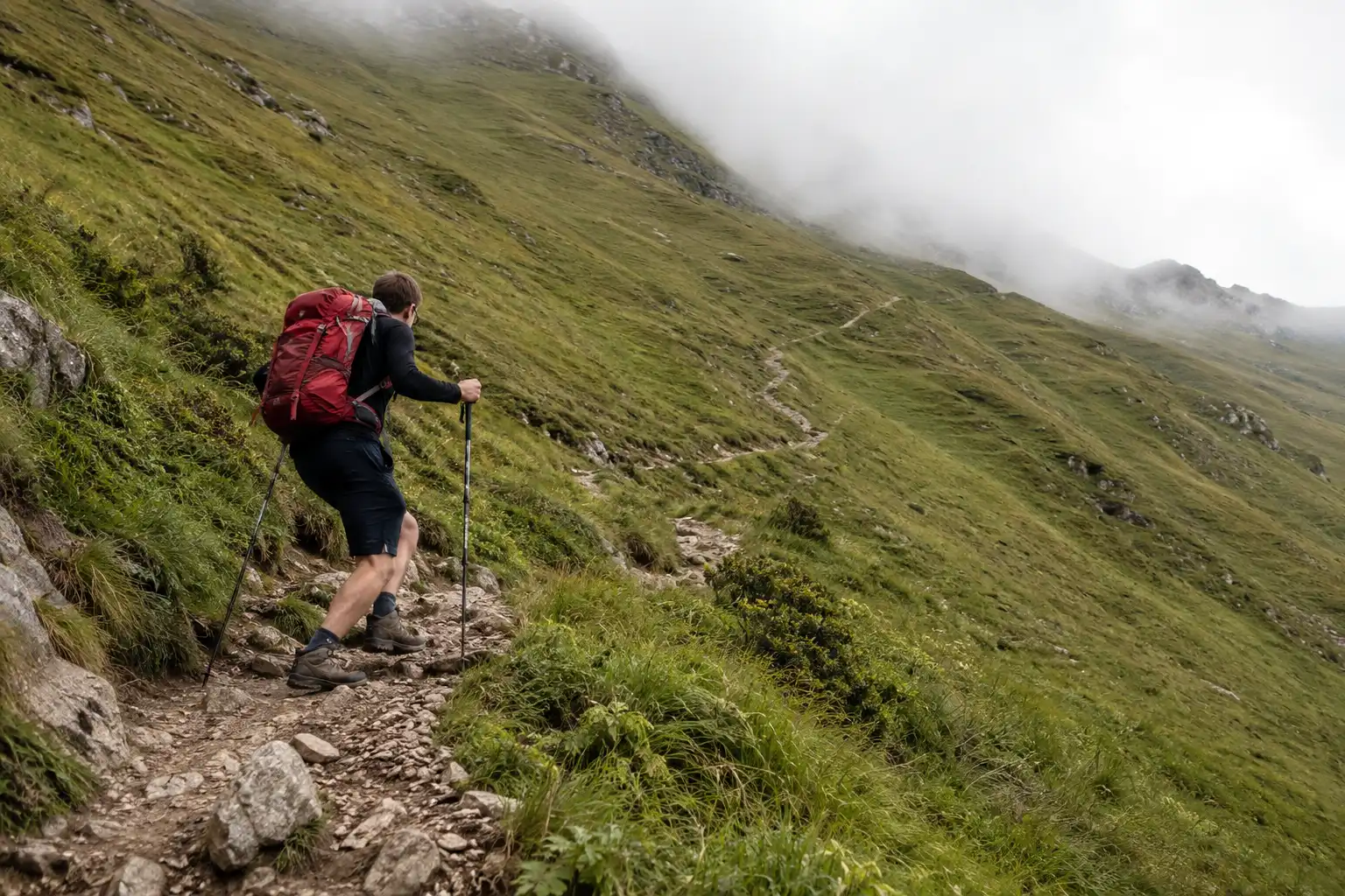 Hiker climbing a steep switchback section of trail with trekking poles on a grassy hillside