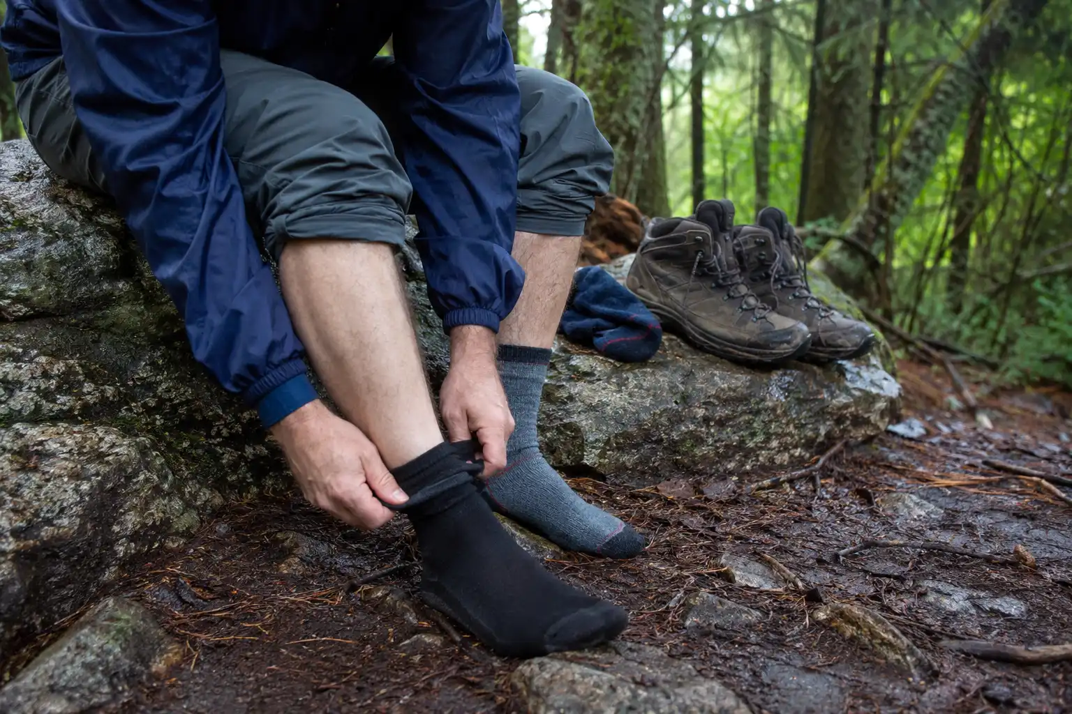 Hiker sitting on a rock pulling a thin liner sock onto one foot with a cushioned outer sock and boot ready beside them