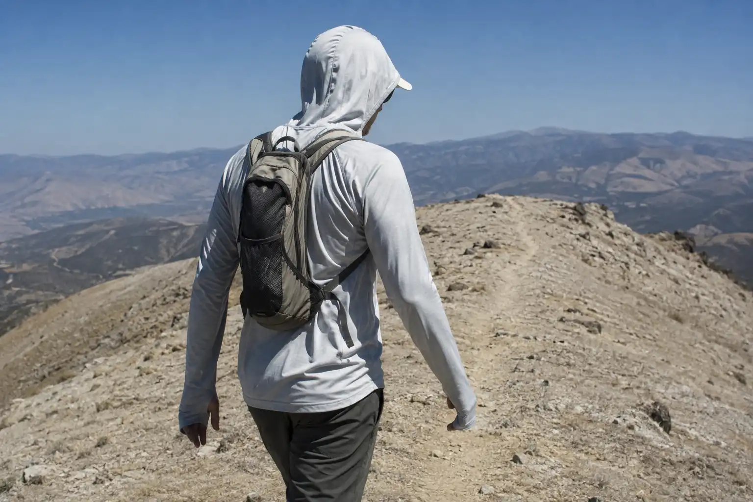 Hiker in a loose light colored long sleeve sun hoodie with the hood raised over a cap, walking an exposed trail in strong sunlight