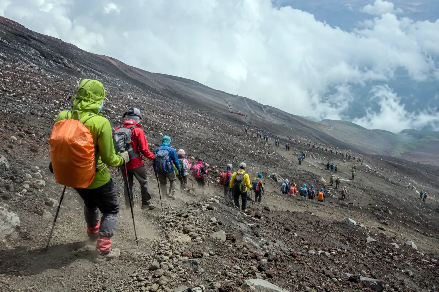 Line of hikers walking down a wide slope of loose gray volcanic gravel on Mount Fuji with dust rising around their boots