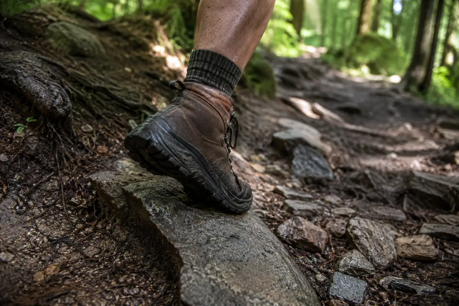 Close view of a hiking boot planted on loose rock and exposed tree roots with the ankle angled to hold balance on uneven ground