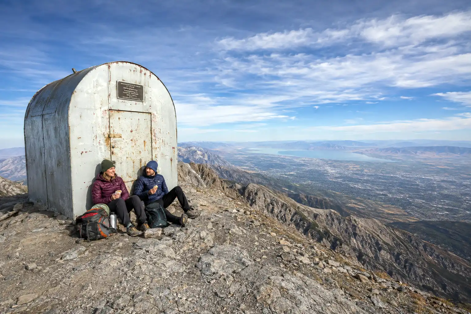 Small metal shelter hut on the summit of Mount Timpanogos with hikers resting beside it and Utah Valley visible far below