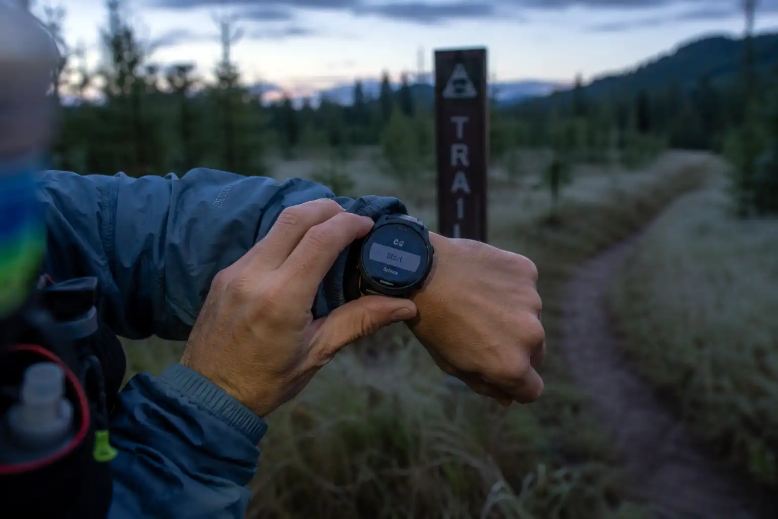 Hiker starting the timer on a GPS watch at a quiet trailhead sign early in the morning with no other people around