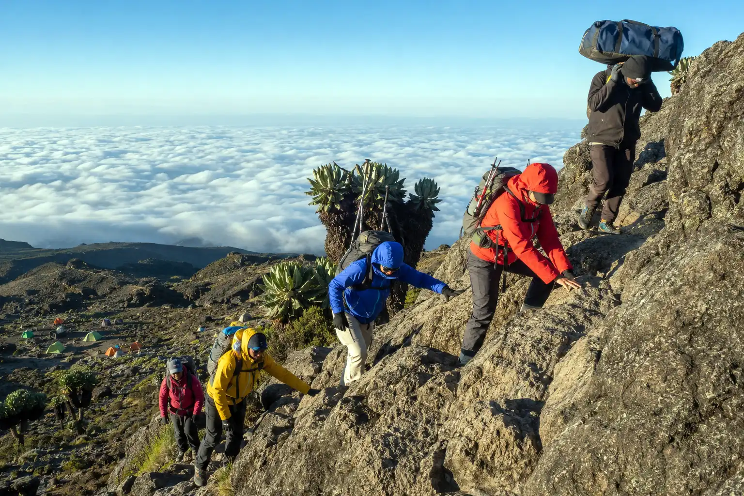 Trekkers using their hands to scramble up a rocky section of the Barranco Wall on Kilimanjaro with poles stowed on their packs