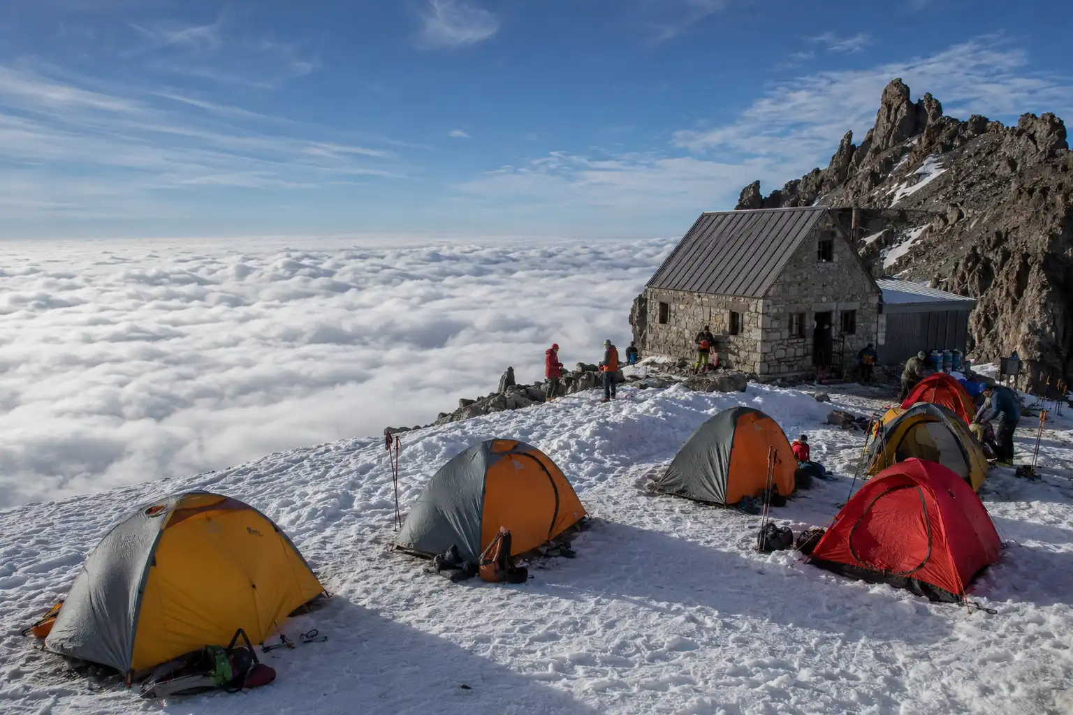 The historic stone shelter at Camp Muir on Mount Rainier surrounded by climber tents pitched on snow with a cloud layer below