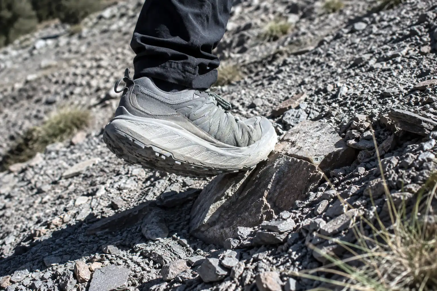 Hiker in a thick cushioned trail shoe stepping onto a loose angled rock on a steep slope, showing reduced stability on technical ground