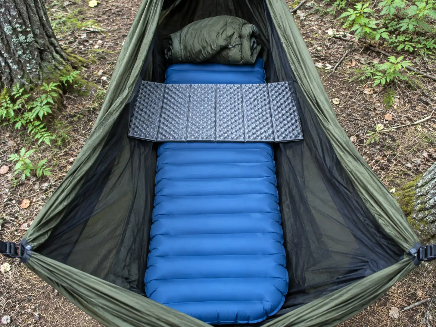 Folded closed cell foam sit pad laid sideways under a camper's shoulders on top of a narrow sleeping pad inside a hammock