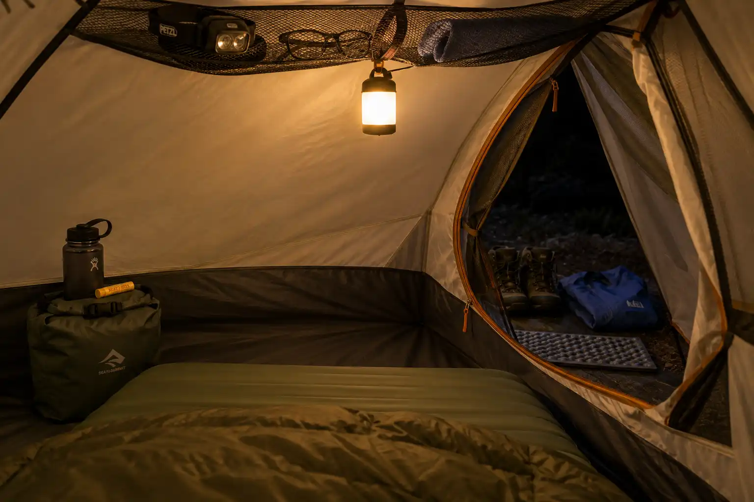 Inside view of a tent at night showing a hanging lantern, a headlamp and glasses in the gear loft, a dry bag beside the sleeping pad, and boots stored in the vestibule