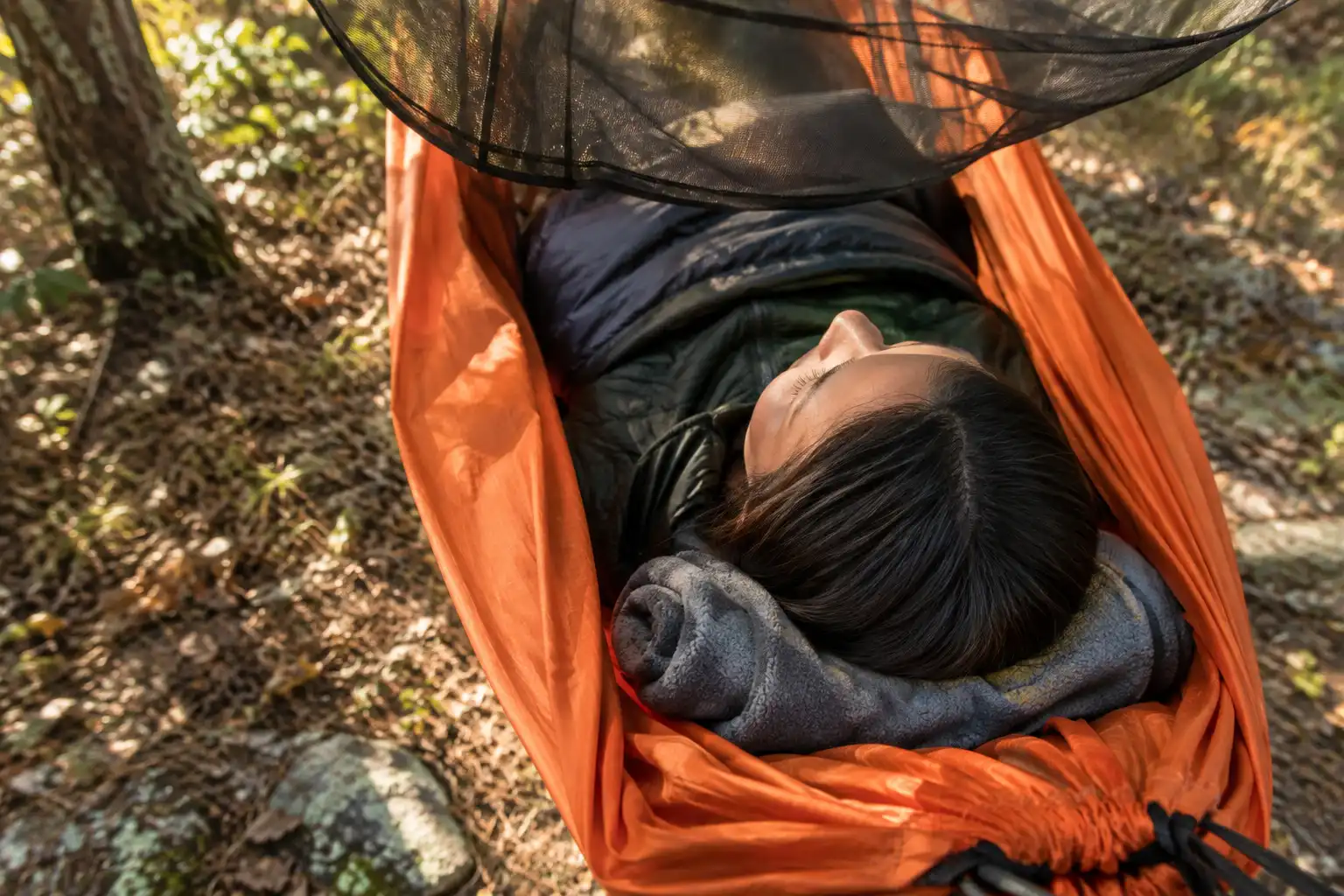 A rolled fleece jacket tucked under a camper's neck at the head end of a camping hammock with no camping pillow in use